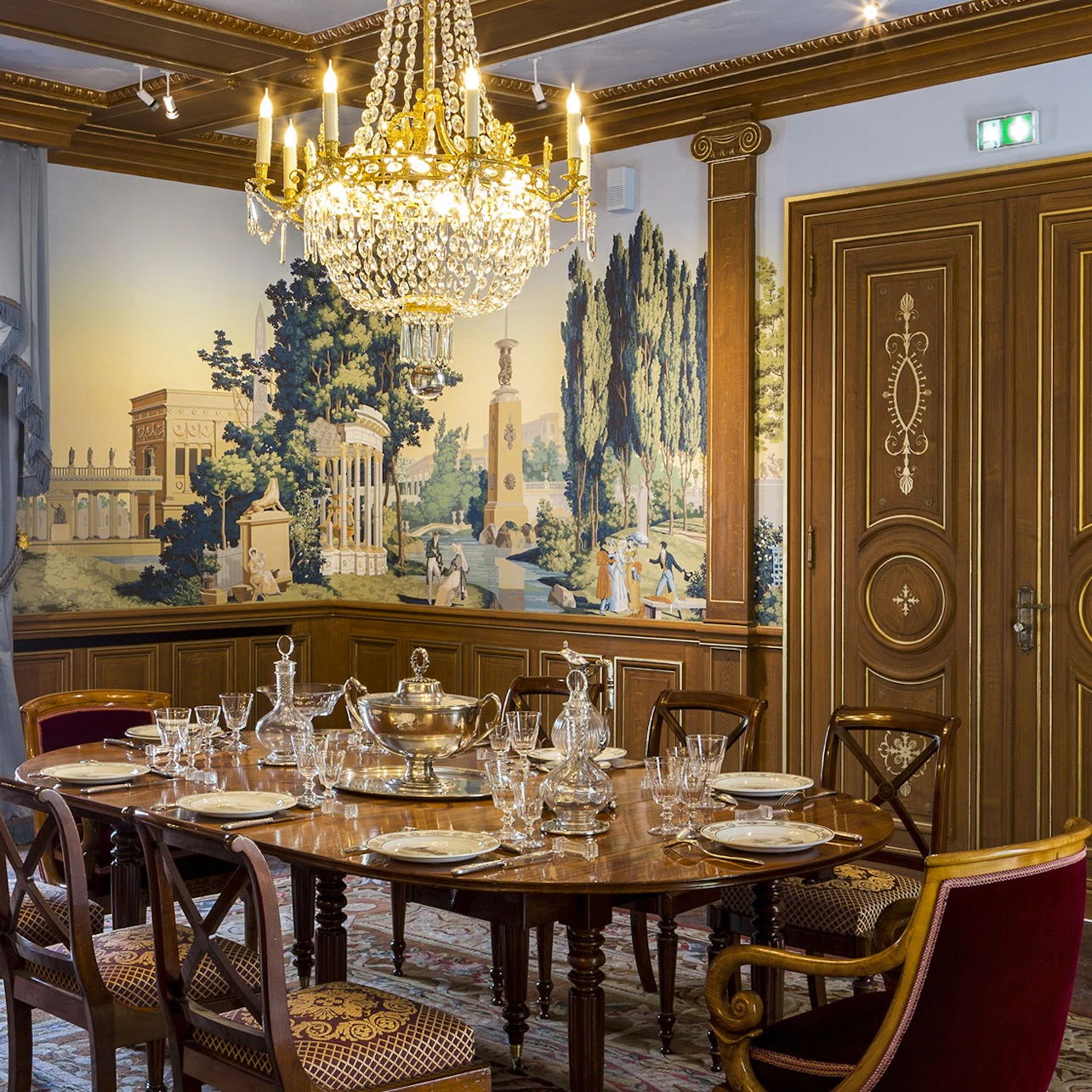 Elegant dining room with a wood table set with dishes and crystal, a chandelier, mural walls, and ornate wooden doors.