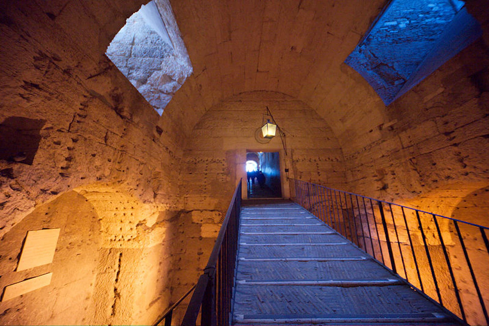 A narrow walkway with railings leads through a stone archway in a dimly lit ancient structure with blue-lit windows.