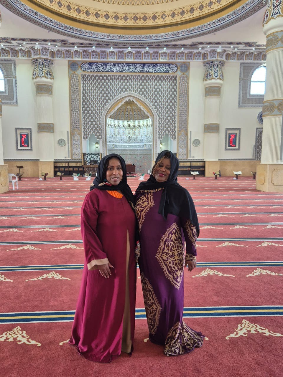 Δύο γυναίκες με σεμνά φορέματα και χιτζάμπ στέκονται μέσα σε ένα περίτεχνο τζαμί με περίπλοκες καμάρες και κόκκινο δάπεδο με χαλιά.