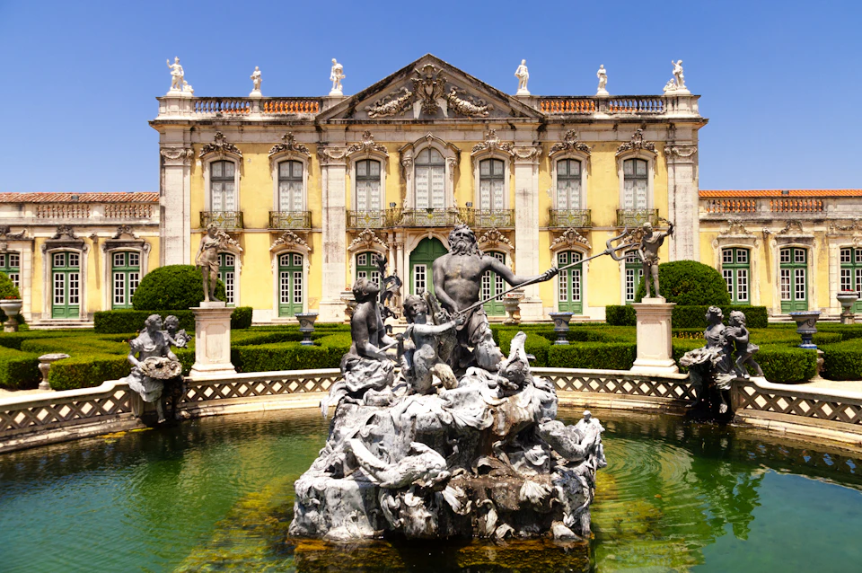 A historic building with ornate details stands behind a fountain featuring numerous marble statues in a manicured garden.