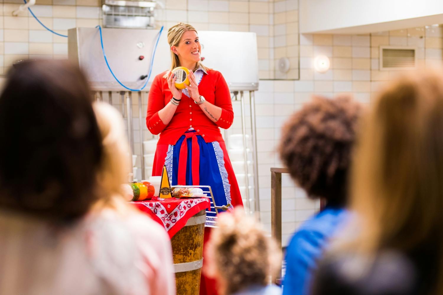 Μια γυναίκα με κόκκινα και μπλε ρούχα κρατάει τυρί ενώ κάνει μια παρουσίαση σε ένα μικρό κοινό σε ένα δωμάτιο με πλακάκια.