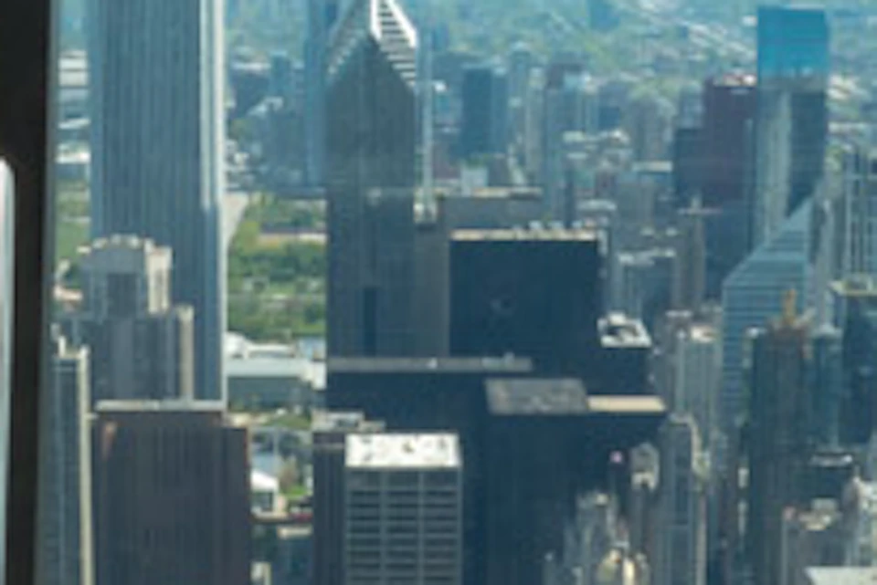 Man and girl stand in a glass observation platform overlooking a cityscape with tall buildings and a blue sky.