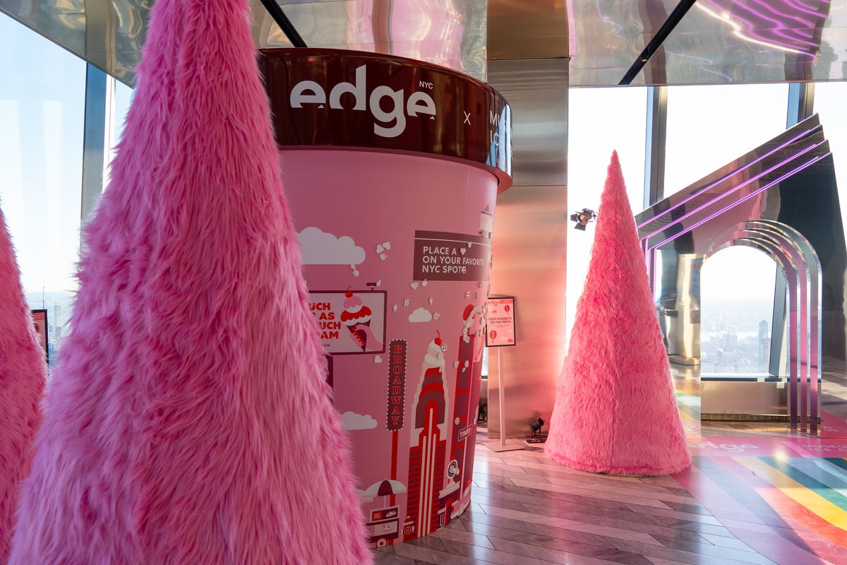 Válcová konstrukce s nápisem „edge“, obklopená dvěma velkými růžovými, chlupatými dekoracemi ve tvaru kužele v moderním interiéru.