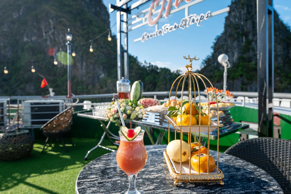 Outdoor dining area with a tropical drink, fruit in a golden cage, buffet table, string lights, and mountain scenery in the background.