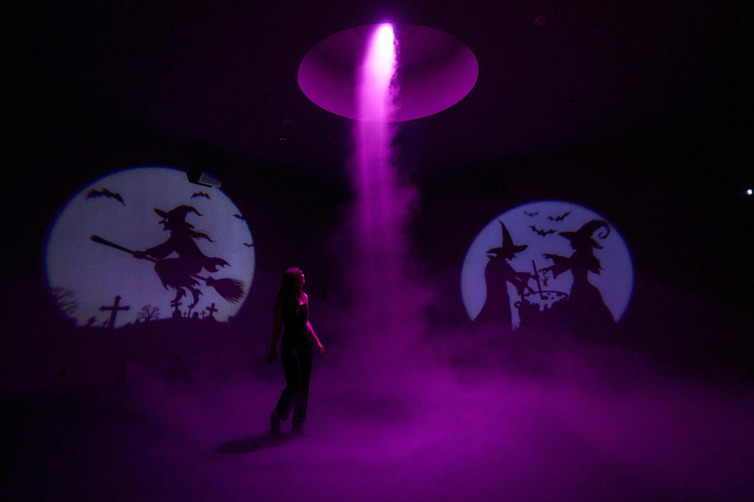 A person stands in a purple-lit room with fog. Witch silhouettes are projected on two circular screens in the background.