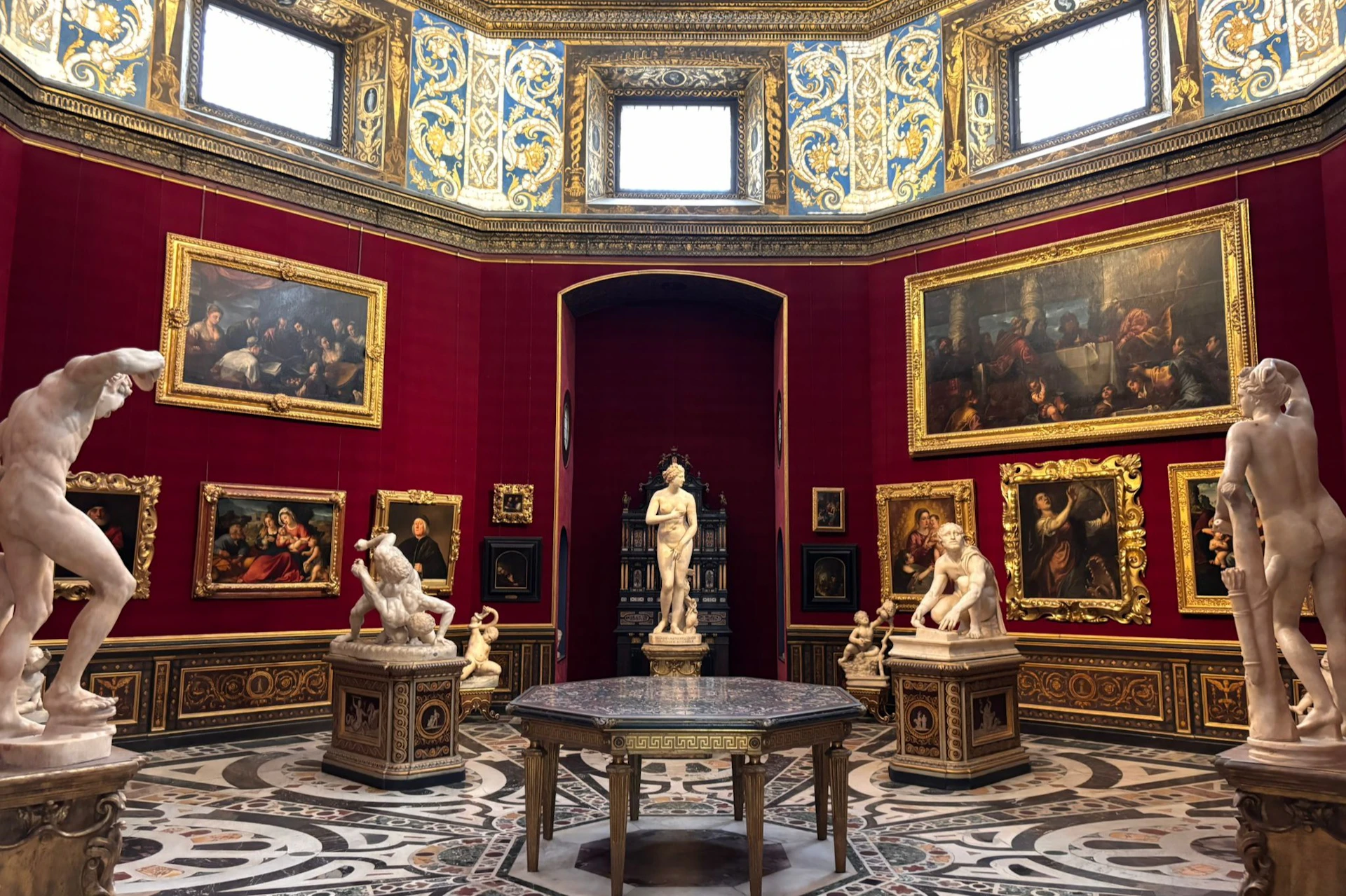 An ornate gallery with red walls, classical sculptures, framed paintings, and a central marble-topped table on a patterned floor.