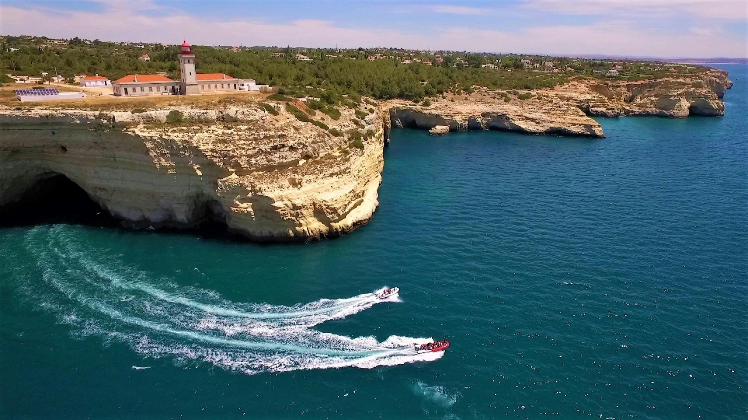 Two speedboats create white trails on blue water near a cliff with a lighthouse and greenery on top.