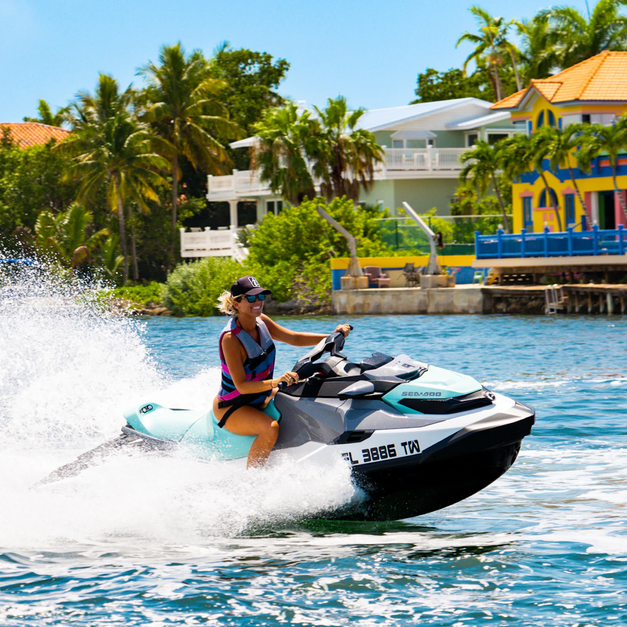 A person riding a jet ski on a sunny day near colorful waterfront houses surrounded by palm trees.