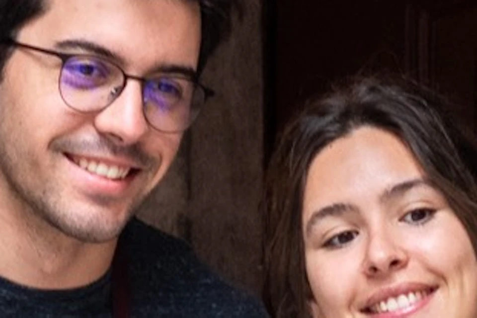Two people smiling and standing by a decorative stone archway with plants and tiled patterns in the background.