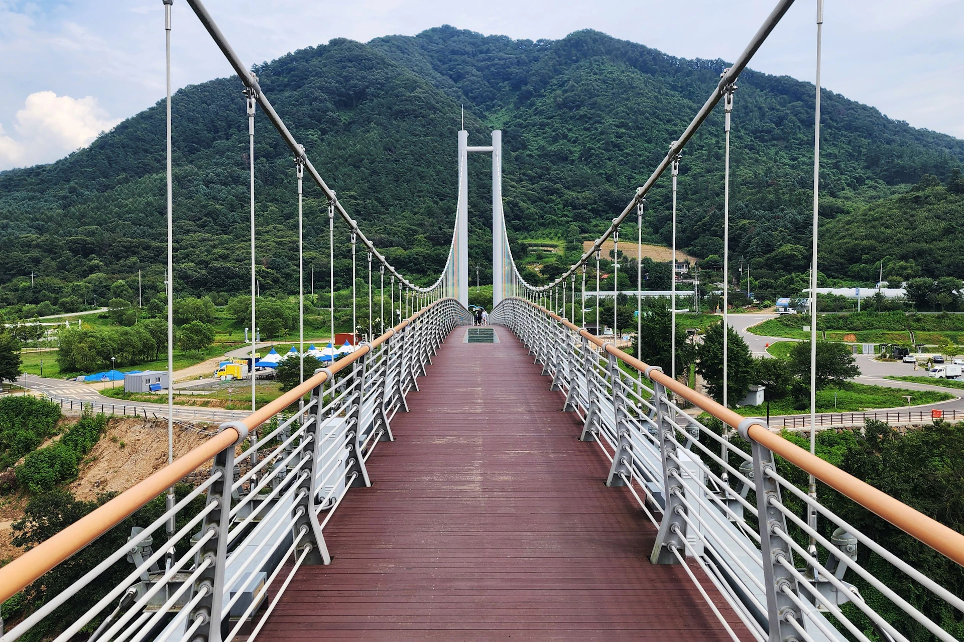 Suspension bridge with wooden decking, white railings, and cable supports, set against a backdrop of lush green mountains.
