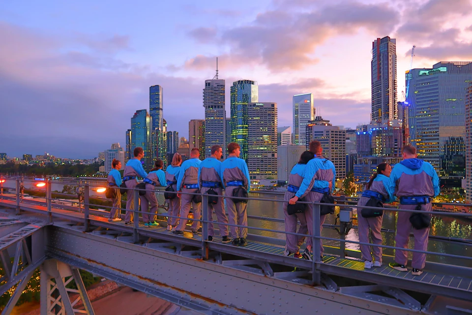 A group of people in blue and gray gear stand on a bridge, overlooking a city skyline at sunset.