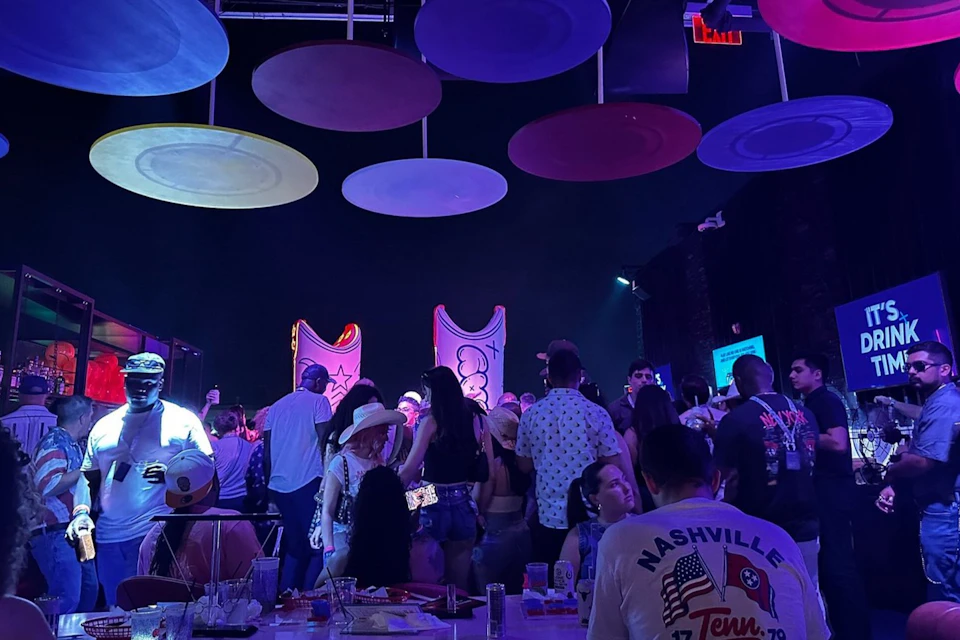 A lively indoor party scene with people dancing under colorful hanging decorations and illuminated artwork in the background.