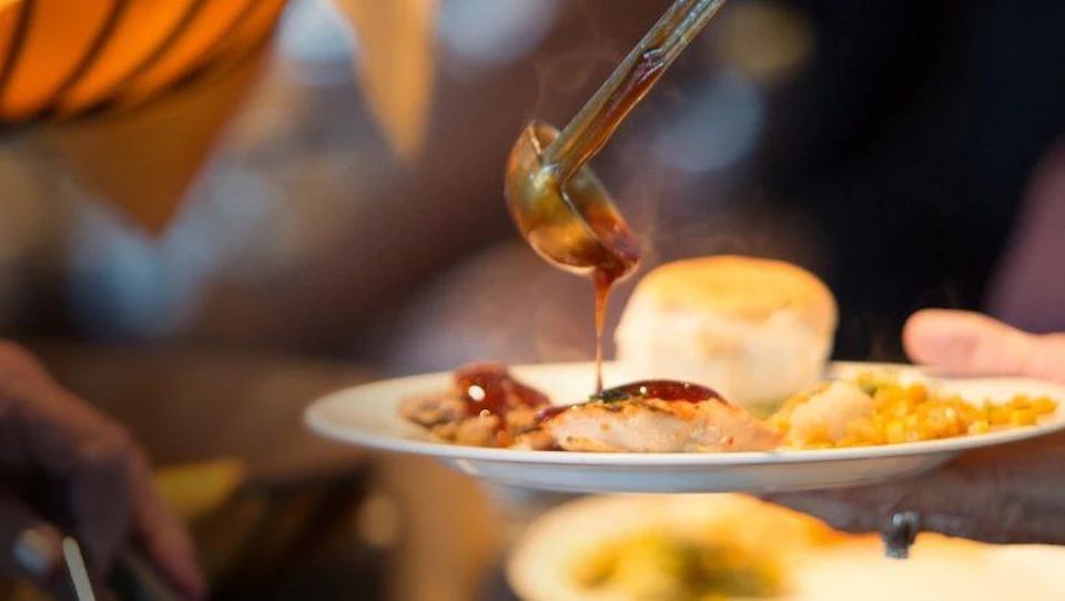 Gravy being ladled onto a plate of food that includes a roll and meat, with steam rising from the dish.