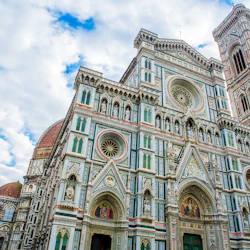 Utsmyckad katedral med invecklad fasad, med cirkulära fönster, statyer och färgglada detaljer mot en delvis molnig himmel.