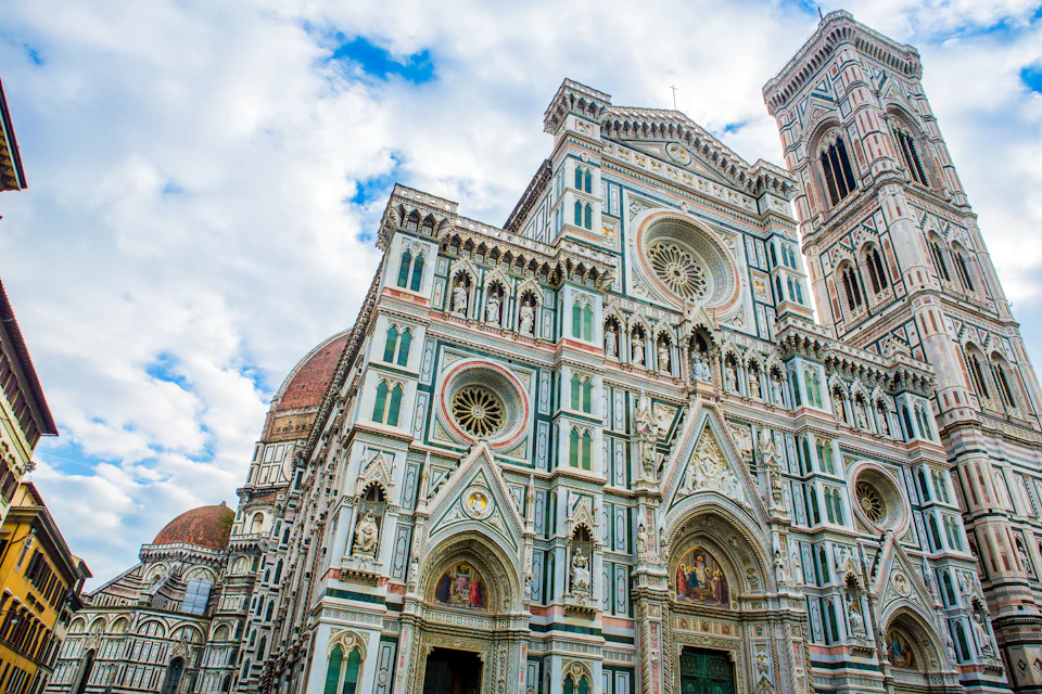 Ornate cathedral with intricate facade, featuring circular windows, statues, and colorful detailing against a partly cloudy sky.