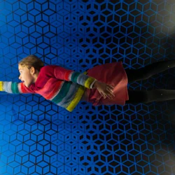 En person i en färgglad tröja verkar flyta i ett futuristiskt rum med en blå geometrisk vägg och glödande paneler på vardera sidan.
