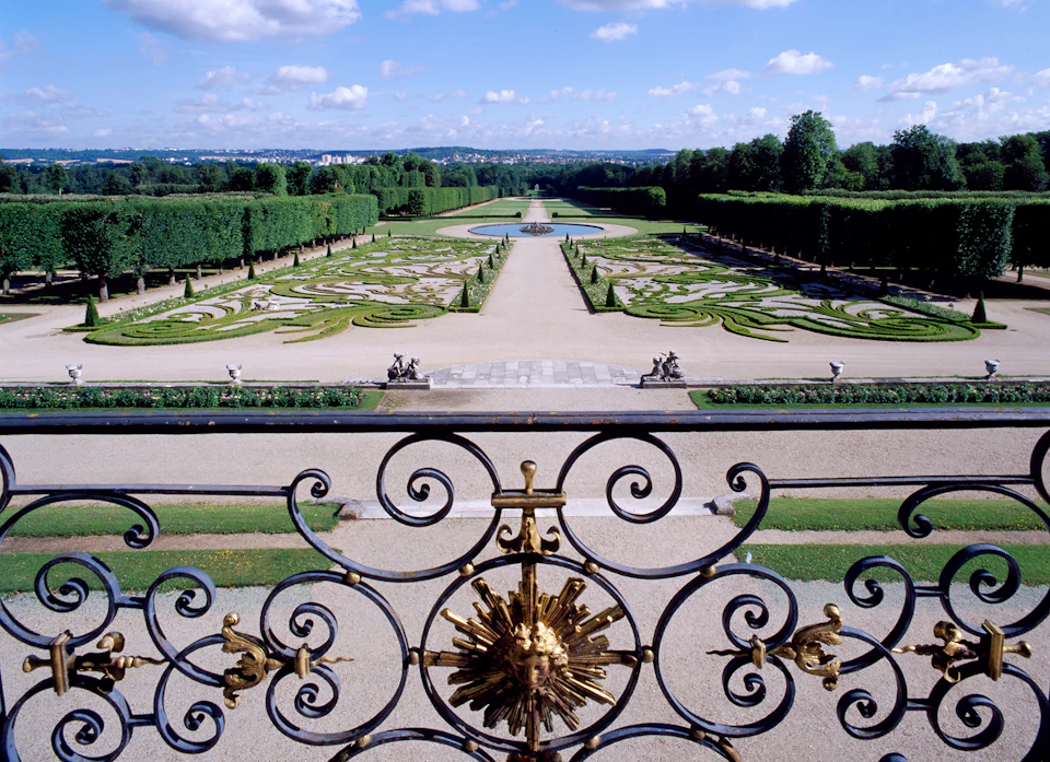 Ornate garden with manicured hedges, pathways, statues, and a central water feature, viewed from behind an intricate wrought iron fence.