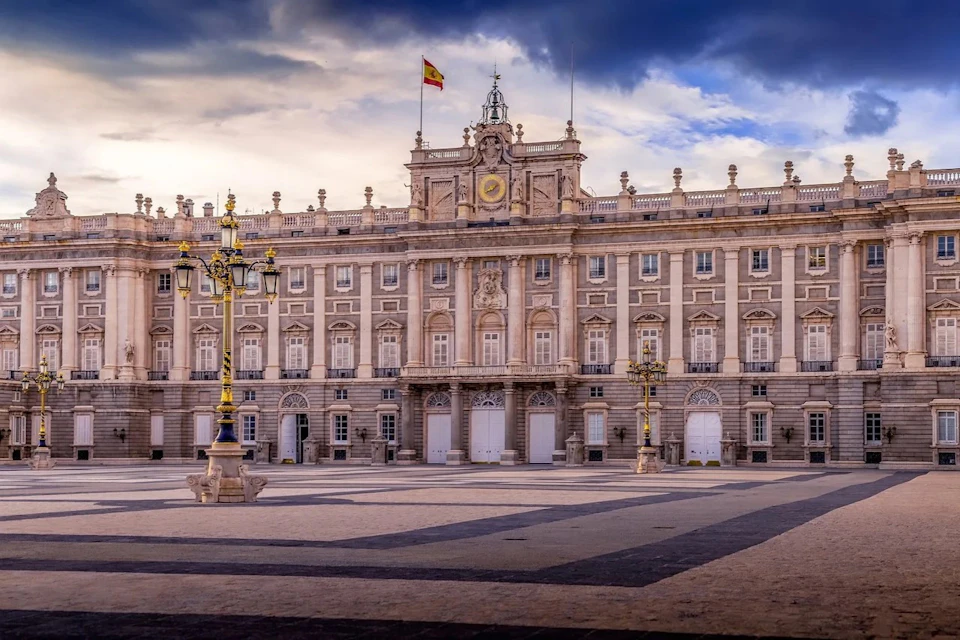 A grand palace with a symmetrical facade, a central clock, and a Spanish flag on top, viewed from an empty courtyard.