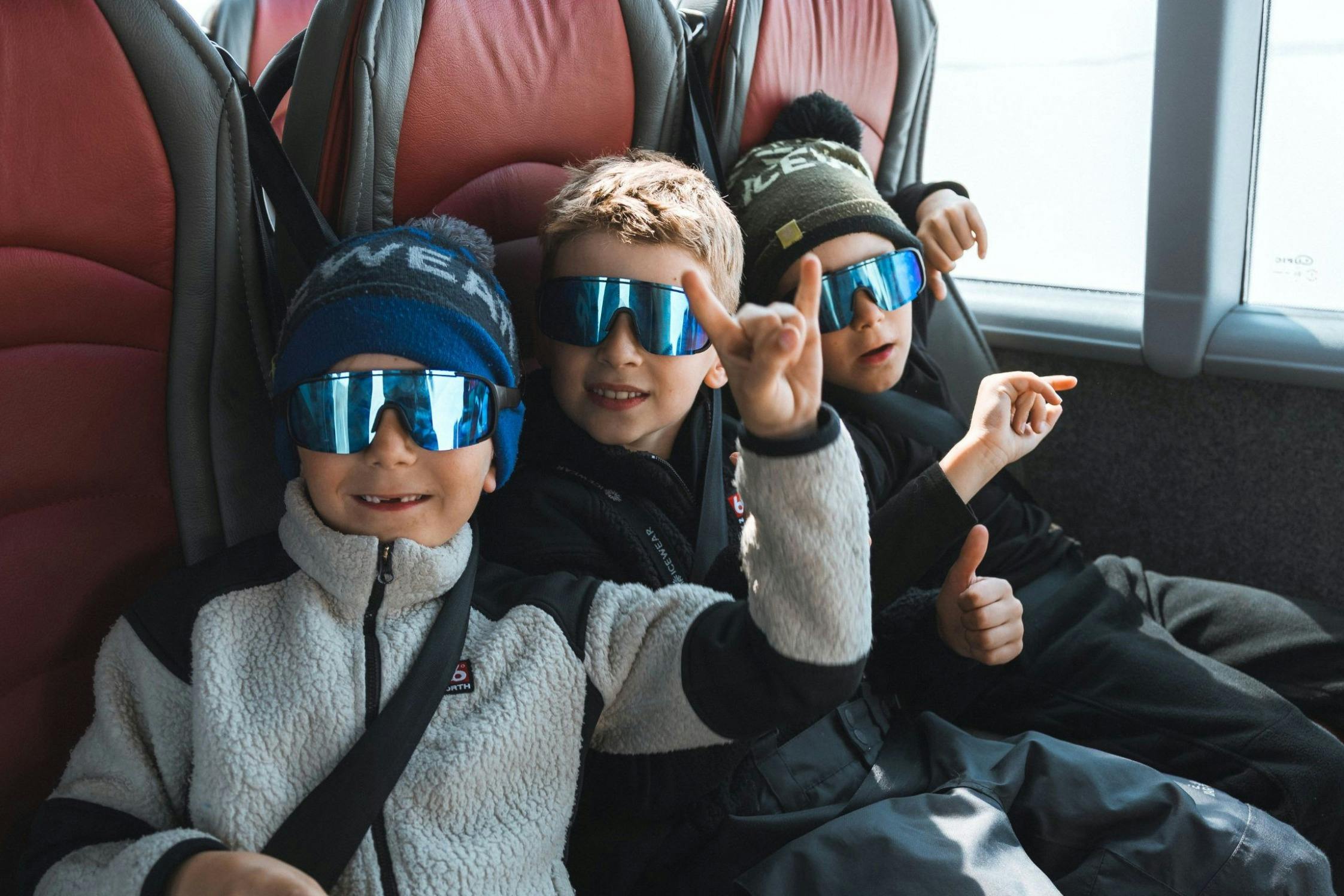 Τρία παιδιά φορώντας γυαλιά ηλίου και χειμωνιάτικα καπέλα, κάθονται σε ένα λεωφορείο, χαμογελώντας και κάνοντας χειρονομίες.