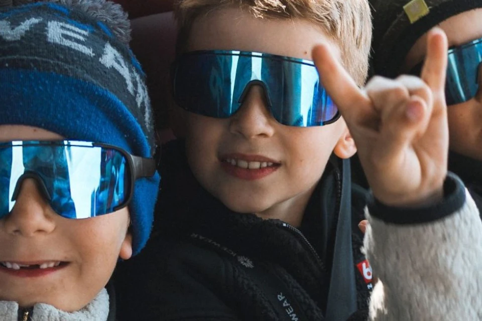 Three children wearing sunglasses and winter hats, seated on a bus, smiling and making hand gestures.