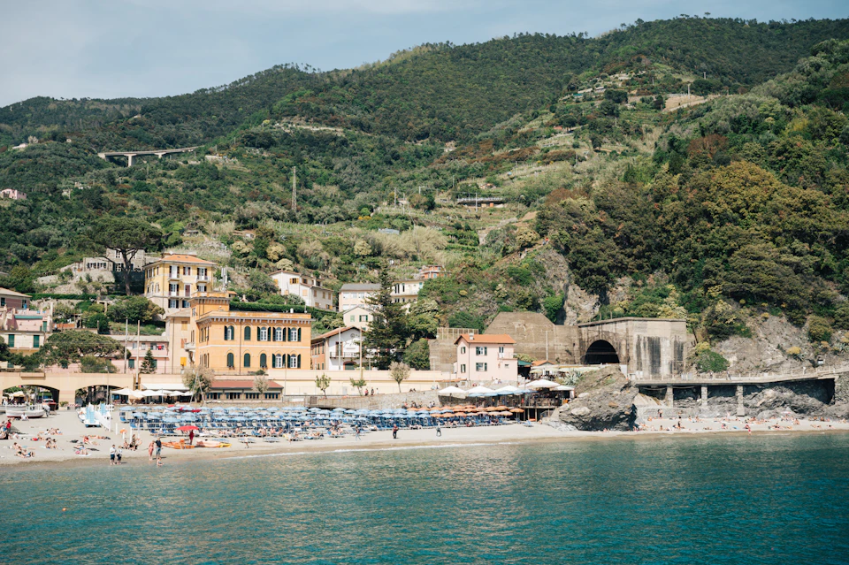 A seaside town with colorful buildings, beach umbrellas, and loungers. Hills with greenery in the background.
