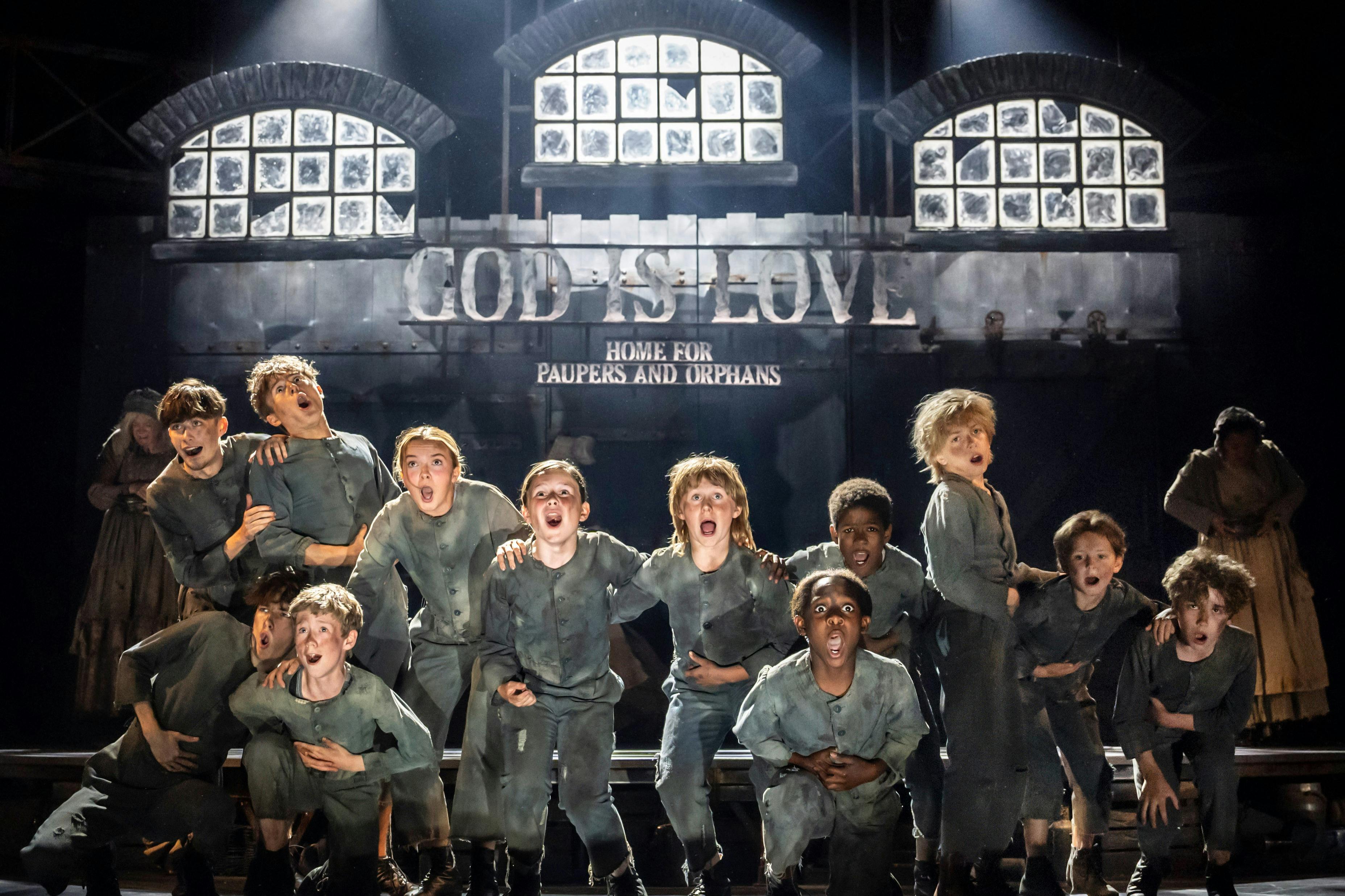 Un grup de nens vestits amb roba andrajosa canten a l'escenari sota un cartell que diu "L'AMOR de DÉUés CASA PER A POBRES I ORFES".