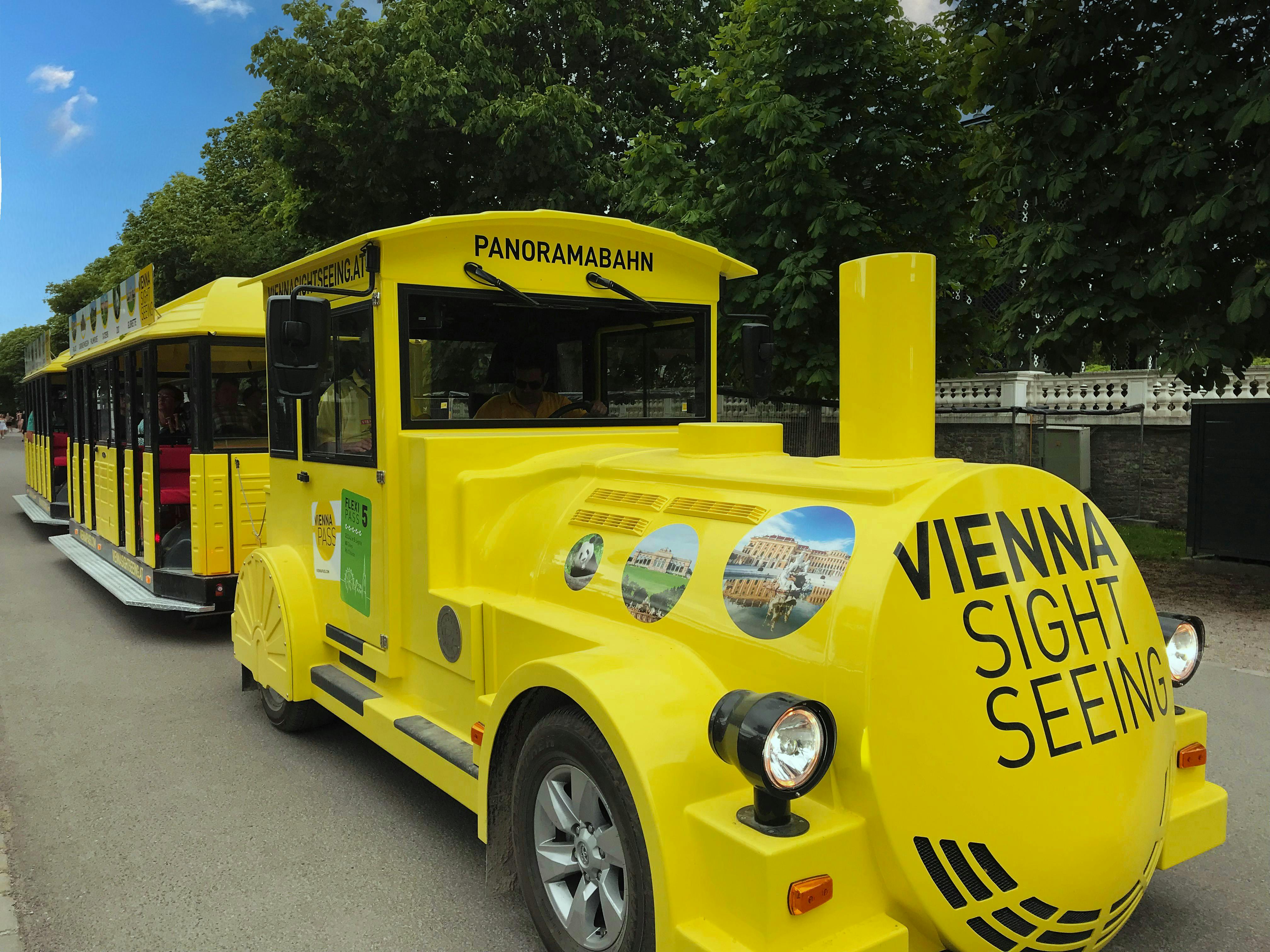 Ett ljust gult sightseeingtåg märkt "Vienna Sightseeing Tours" med träd och en klar himmel i bakgrunden.
