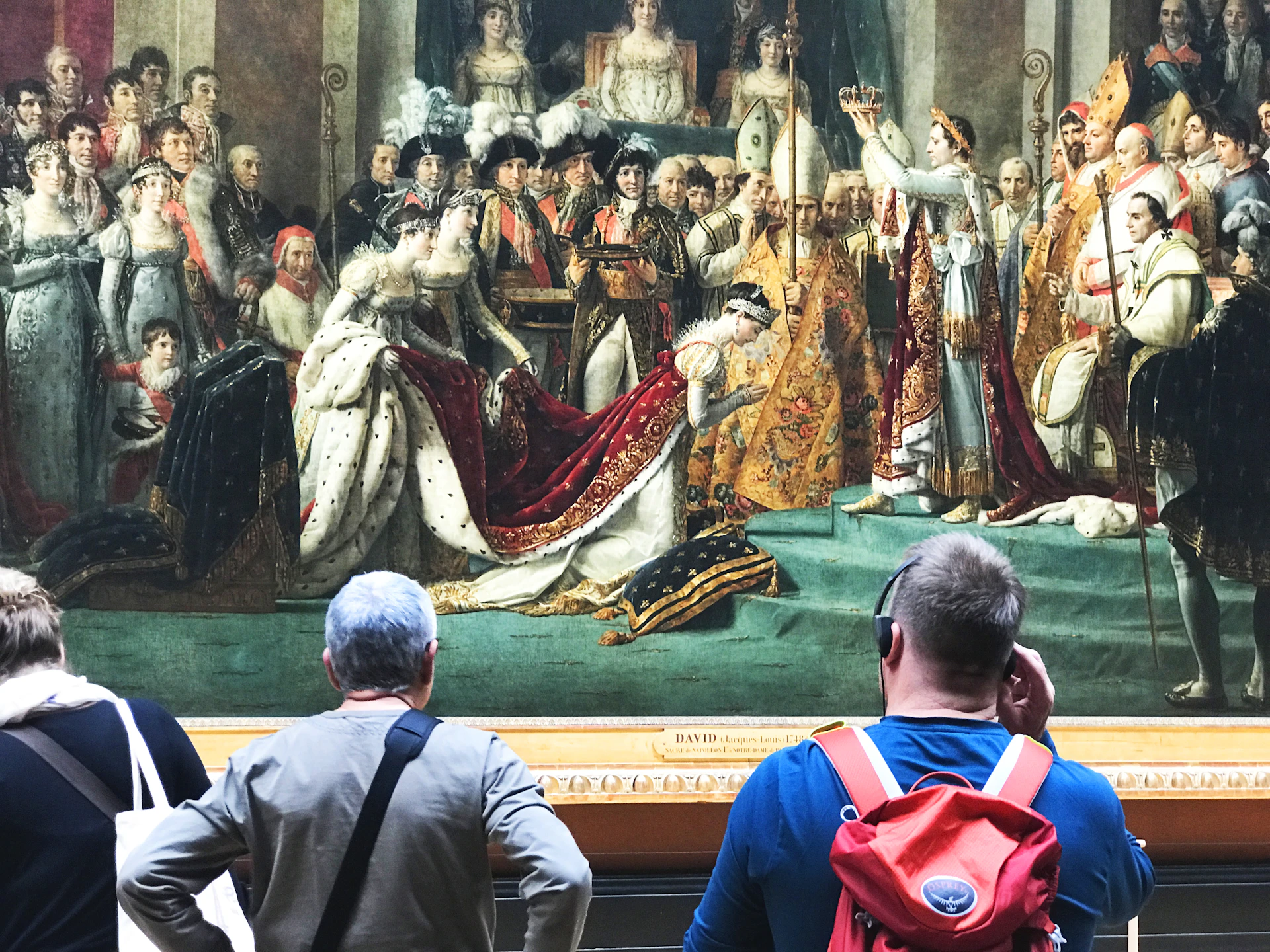 Two people viewing a large historical painting depicting a coronation scene with various figures in opulent attire.