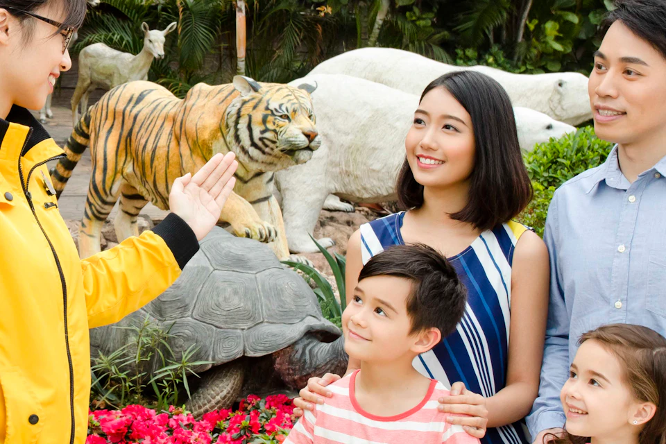 A guide in a yellow jacket talks to a smiling family of four near life-sized animal sculptures outside an attraction.