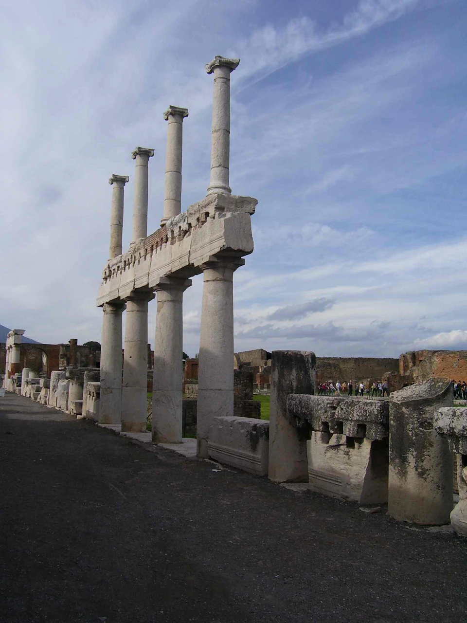 Ancient pillars and ruins stand under a cloudy sky, with stone remnants on the ground, and people visible in the background.