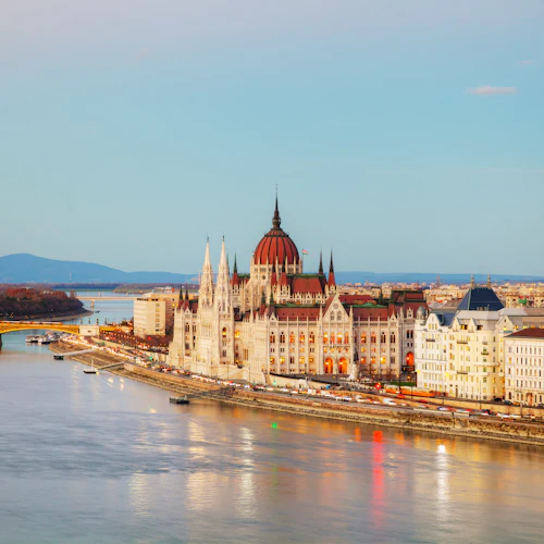 En stadsbild med en stor, utsmyckad byggnad med en kupol och spiror bredvid en flod, med en bro och kullar i bakgrunden.
