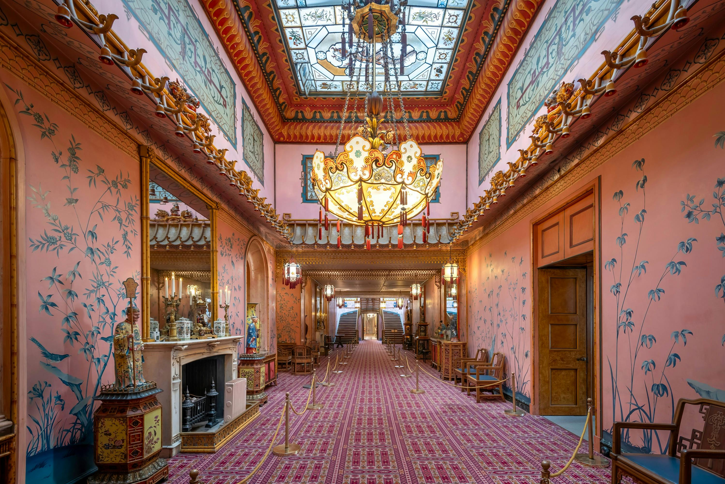 Ornate hallway with a grand chandelier, decorated walls, a large mirror above a fireplace, and a patterned carpet leading to stairs.