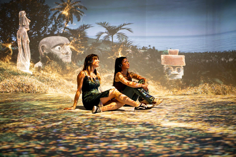 Two women sit on a vividly illuminated floor with a tropical background projection including palm trees and statues.