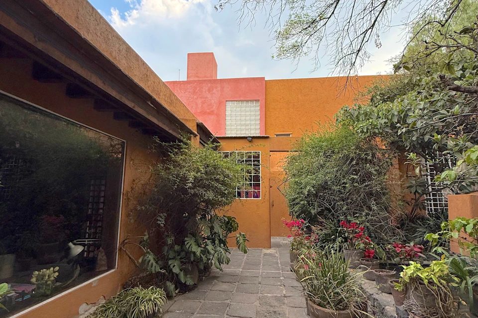 Small courtyard with orange and pink buildings, potted plants, trees, and a stone pathway under a partly cloudy sky.