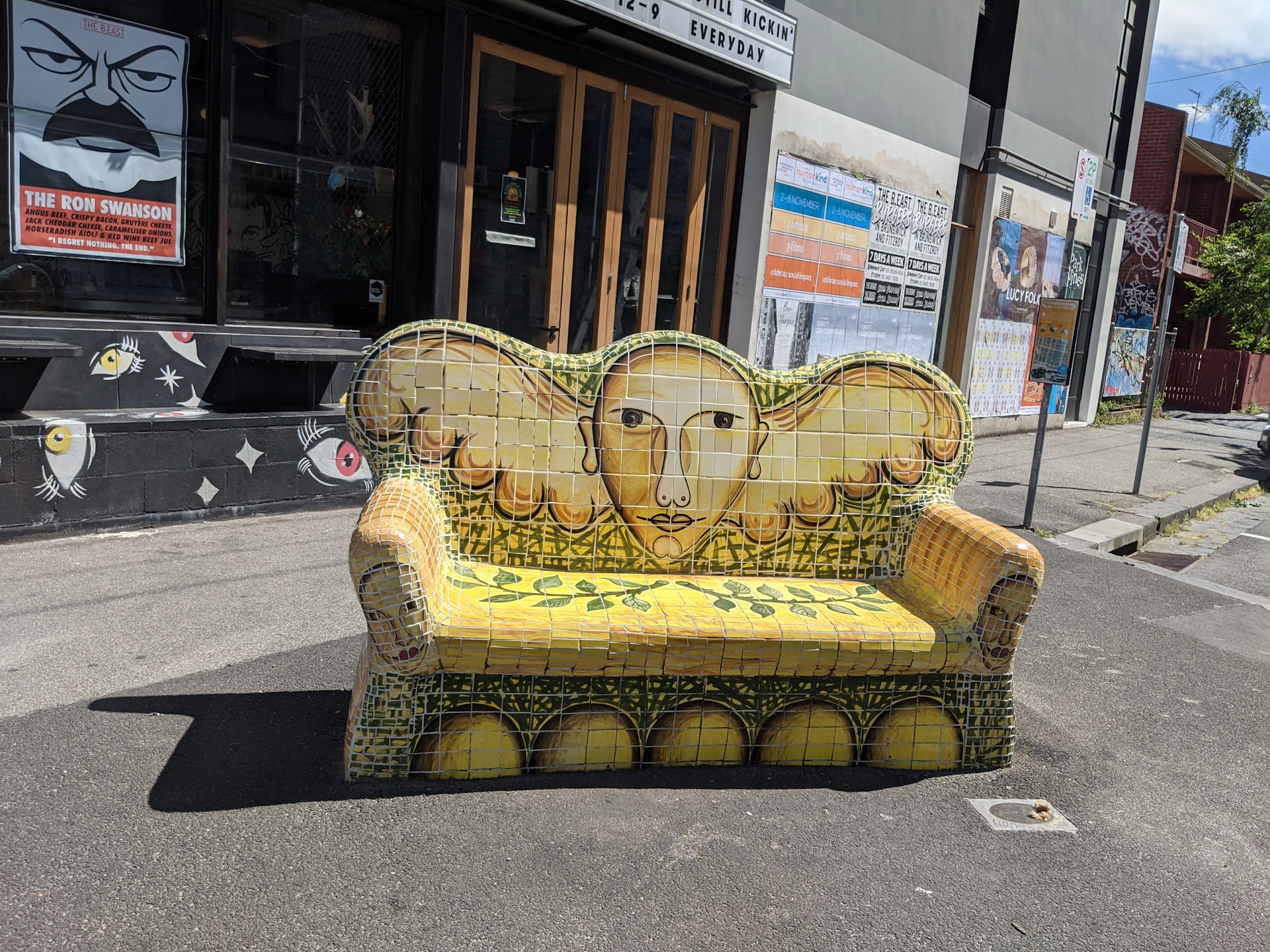 A mosaic bench with a face and wing design is placed on a sidewalk in front of a building with posters and signage.