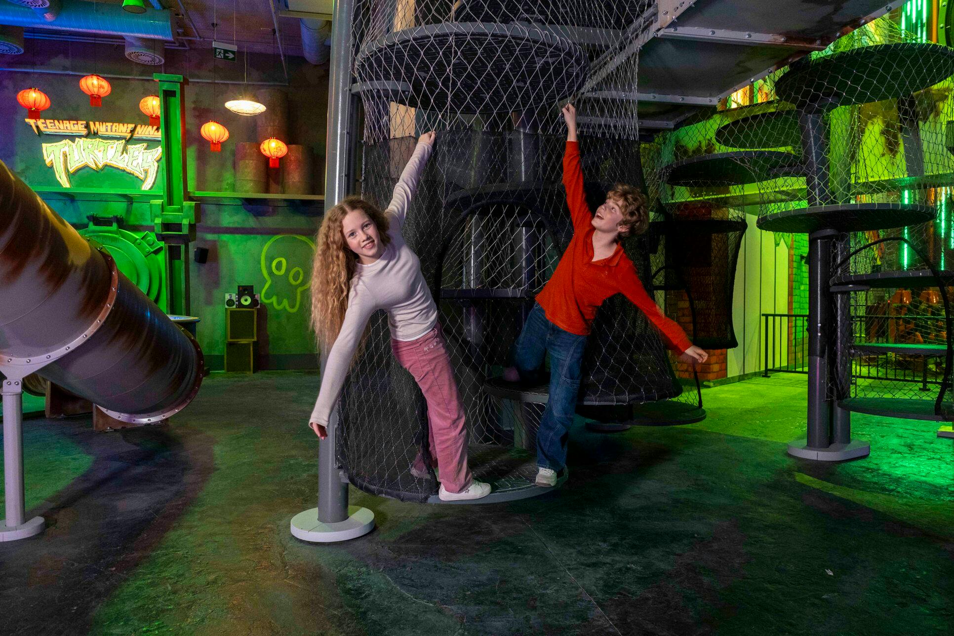 Twee kinderen spelen op een cilindrische netstructuur in een gethematiseerde binnenspeeltuin met kleurrijke lichtjes en decoraties.