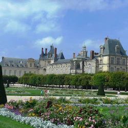 En storslagen historisk byggnad bakom formella trädgårdar med färgglada blommor, geometrisk grönska och vandringsleder under en blå himmel.