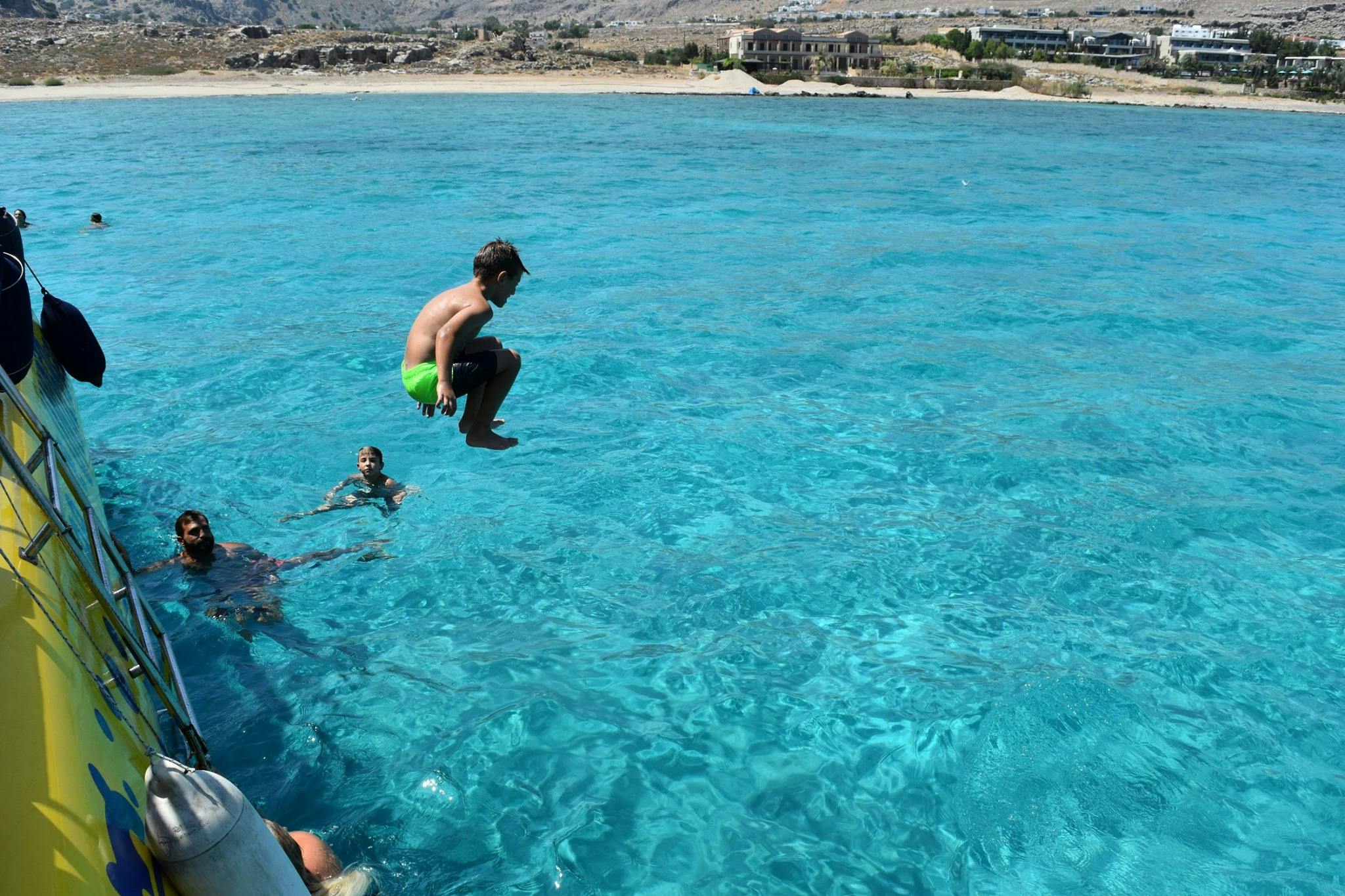 Un ragazzo salta in acque limpide e blu da una barca mentre altre due persone nuotano nelle vicinanze. Sullo sfondo è visibile un paesaggio costiero.