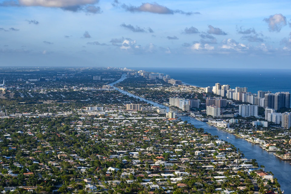 Aerial view of a coastal city with residential areas, high-rise buildings, and a waterway leading to the ocean under a partly cloudy sky.