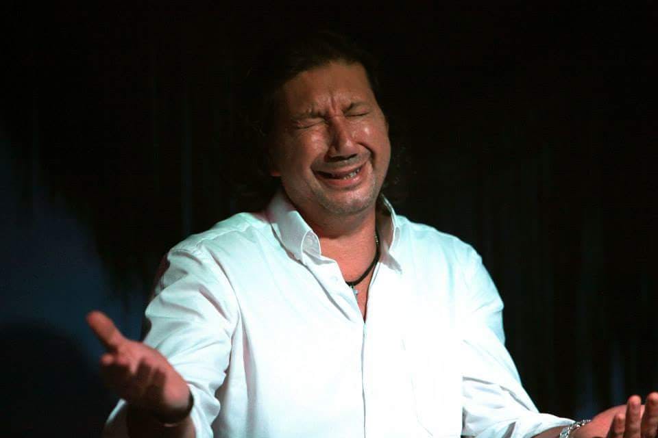 A man in a white shirt with closed eyes and an expressive, emotional facial expression in a dimly lit setting.