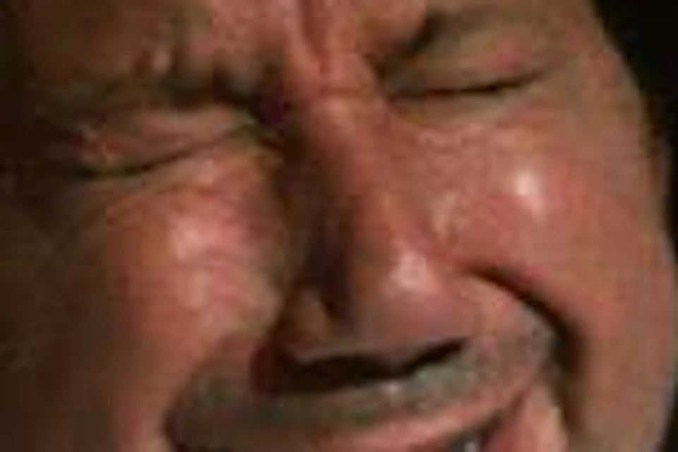 A man in a white shirt with closed eyes and an expressive, emotional facial expression in a dimly lit setting.