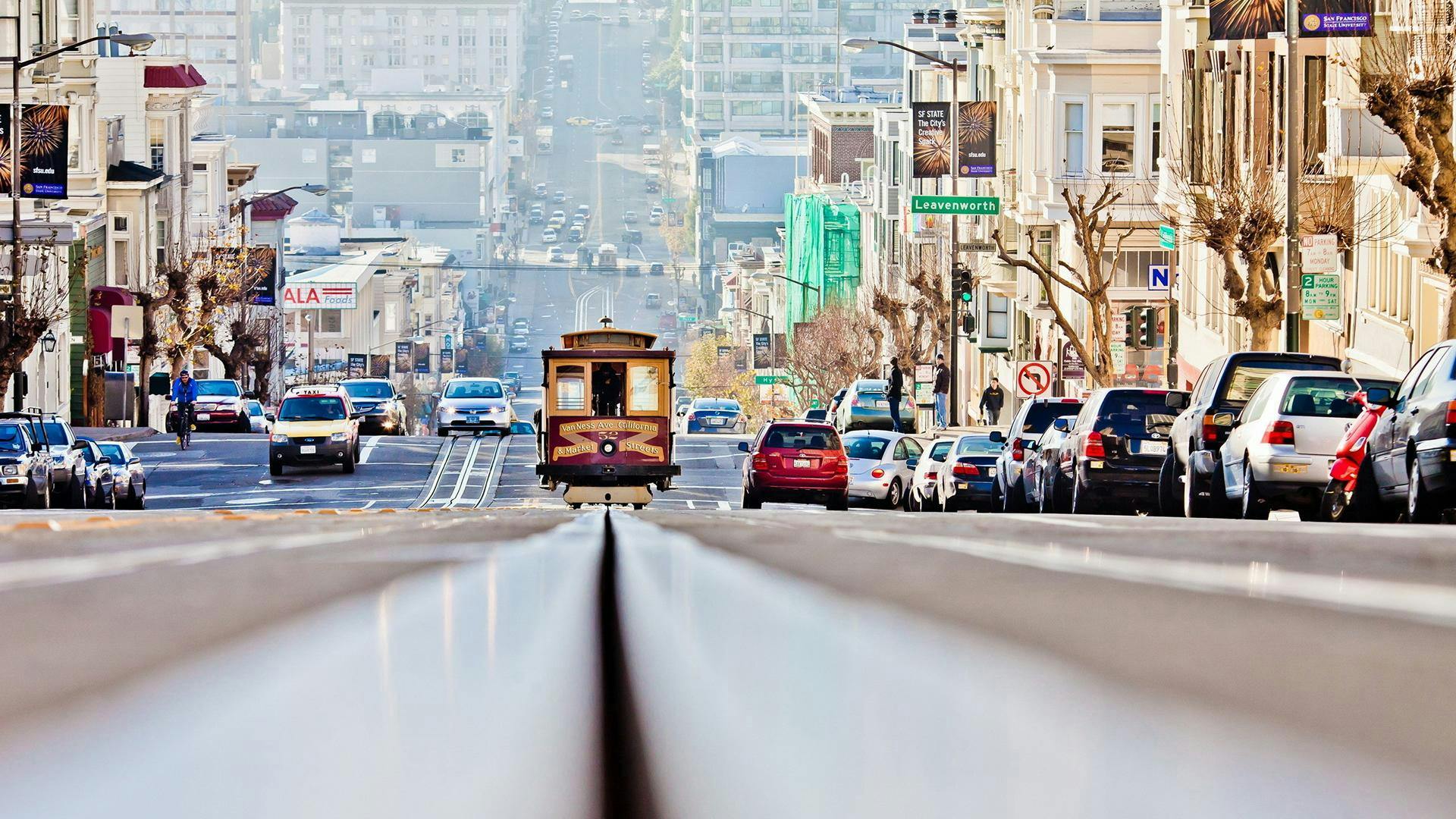 Un tramway monte une rue animée, bordée de voitures et d'immeubles, avec un panneau "Leavenworth" visible.