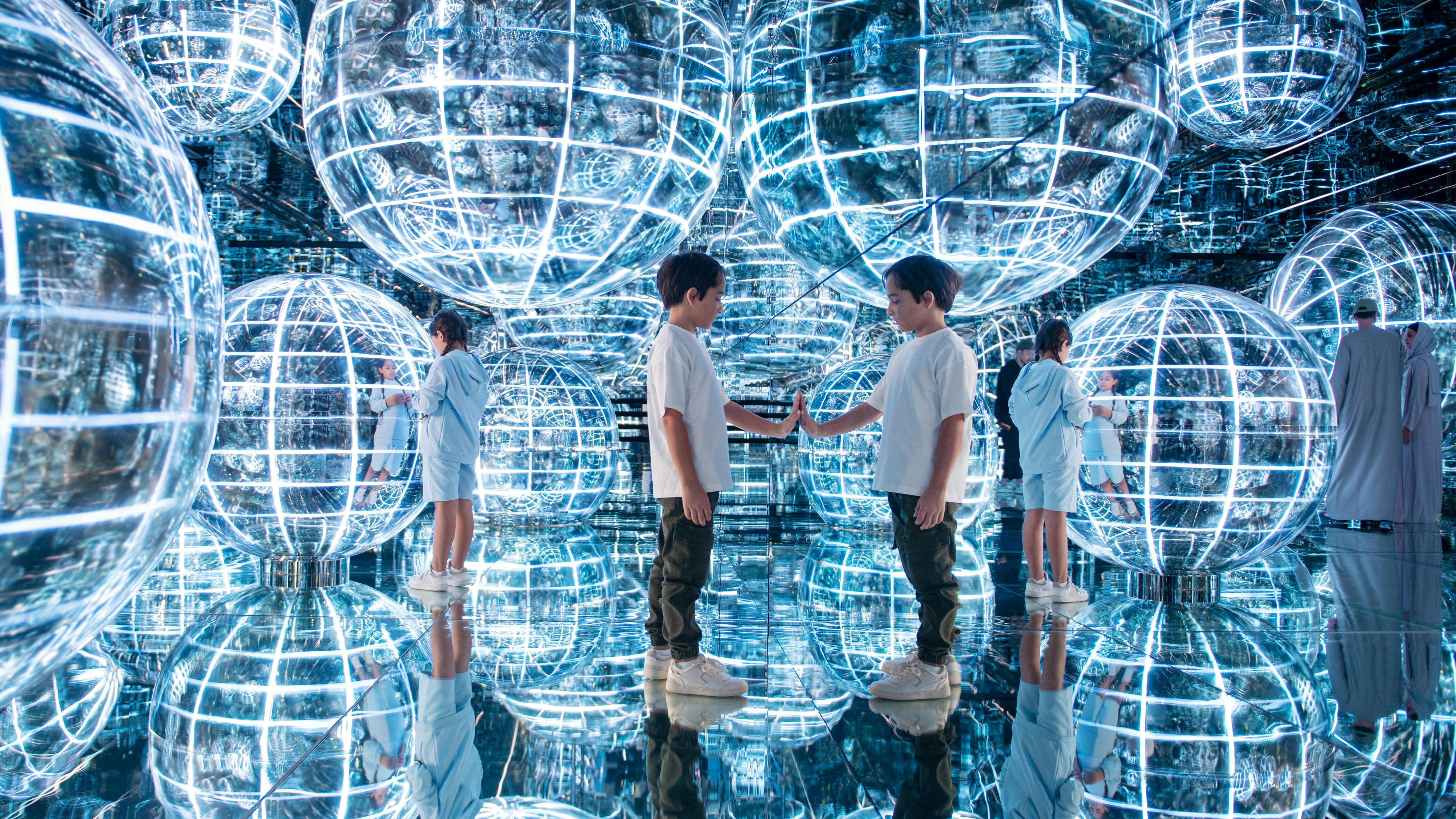 Τα παιδιά αλληλεπιδρούν με κατοπτρικές επιφάνειες και μεγάλες λαμπερές σφαίρες σε έναν φουτουριστικό, ανακλαστικό χώρο.