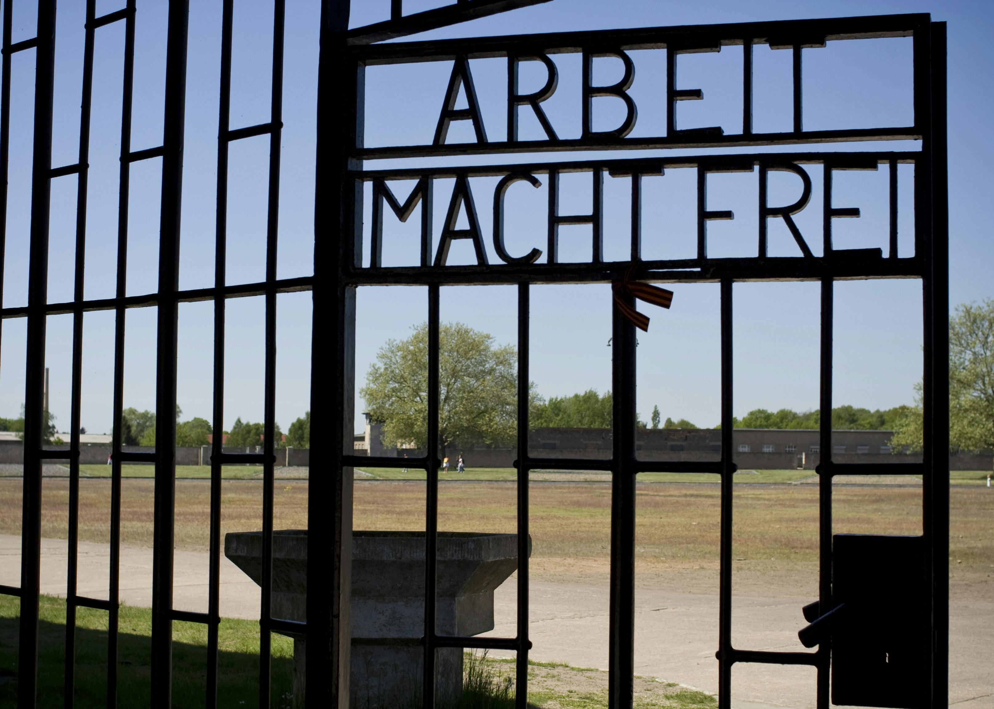 Un portail métallique portant l&#39;inscription &#34;Arbeit macht frei&#34; surplombe un champ stérile et des arbres sous un ciel bleu clair.