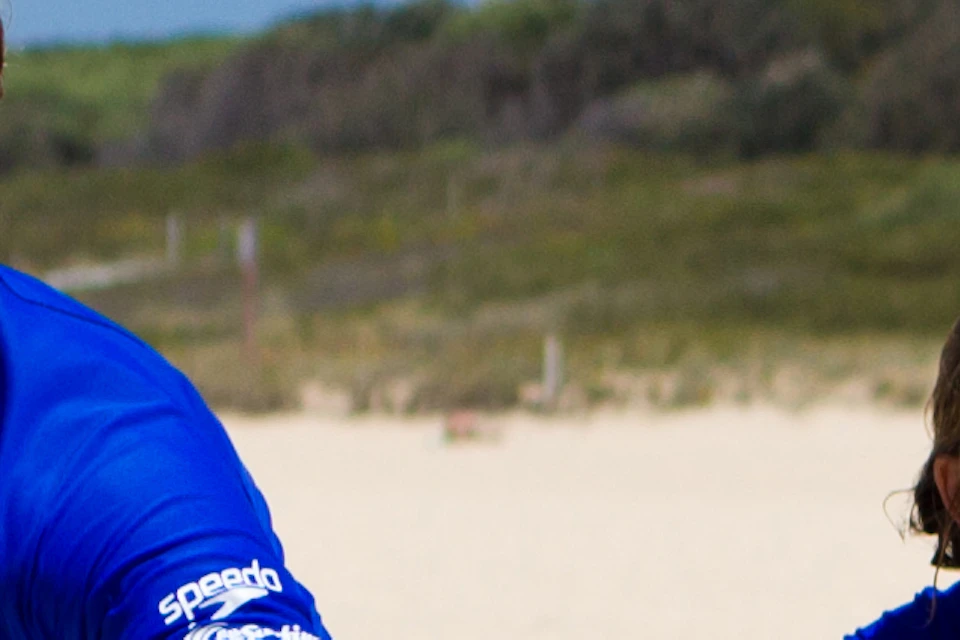 Two people in blue shirts practice surfing stances on blue surfboards on a sandy beach under a clear blue sky.