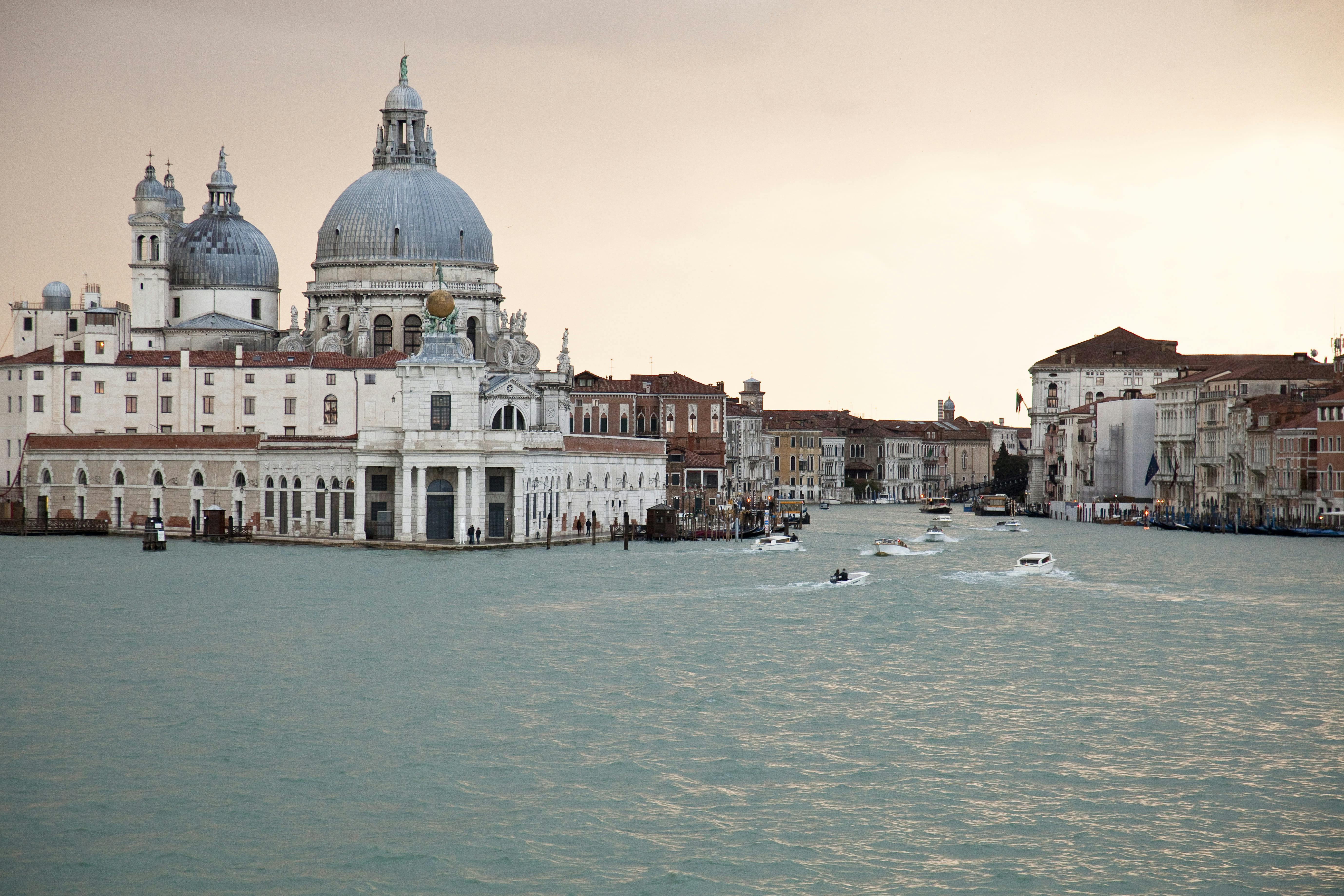 En stor välvd byggnad och andra historiska strukturer vid en kanal med flera båtar i Venedig under en mulen himmel.
