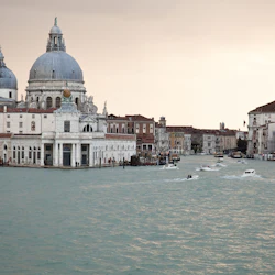 En stor välvd byggnad och andra historiska strukturer vid en kanal med flera båtar i Venedig under en mulen himmel.