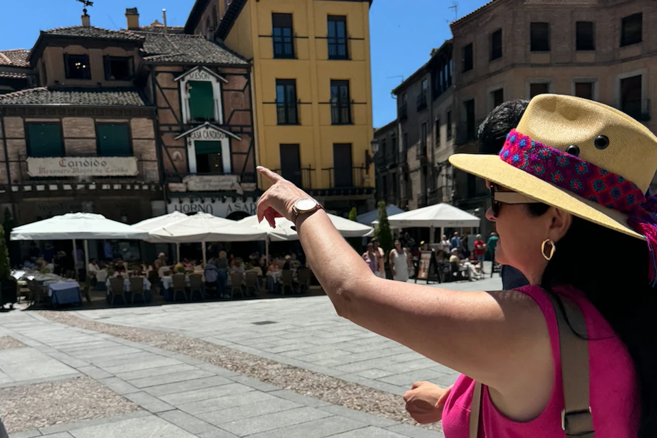 A person wearing a hat and pink top points towards a plaza with outdoor seating and historic buildings in the background.