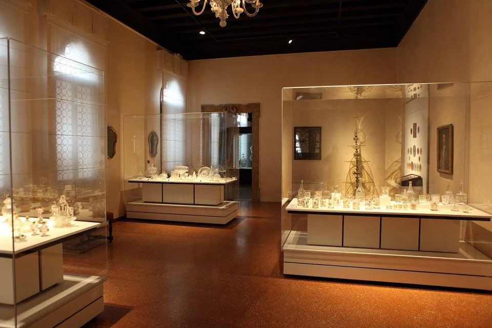 A museum room with glass display cases containing various artifacts, framed artwork on the walls, and a chandelier on the ceiling.
