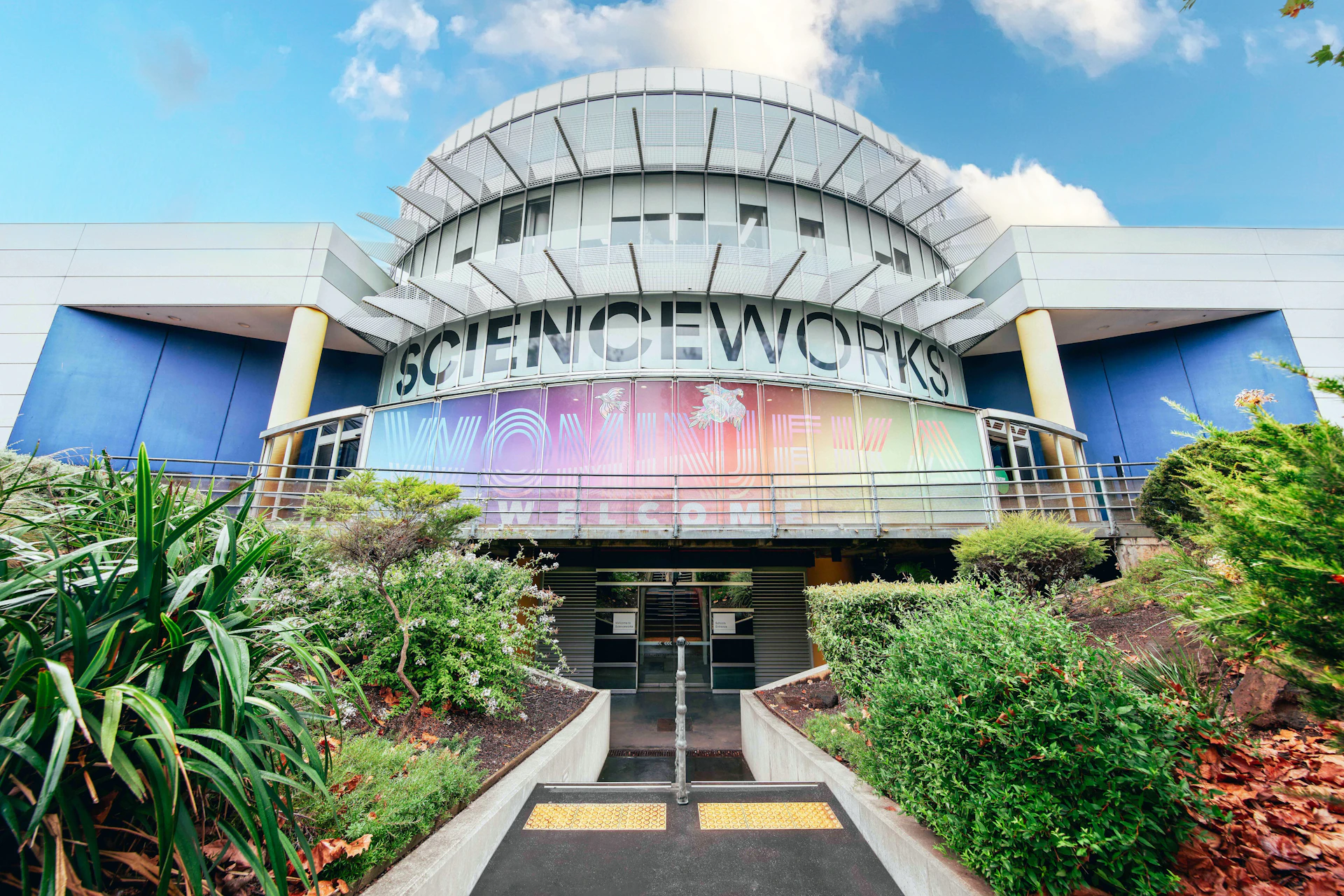Exterior of Scienceworks Museum with colorful signage, modern architecture, and landscaped greenery under a blue sky.